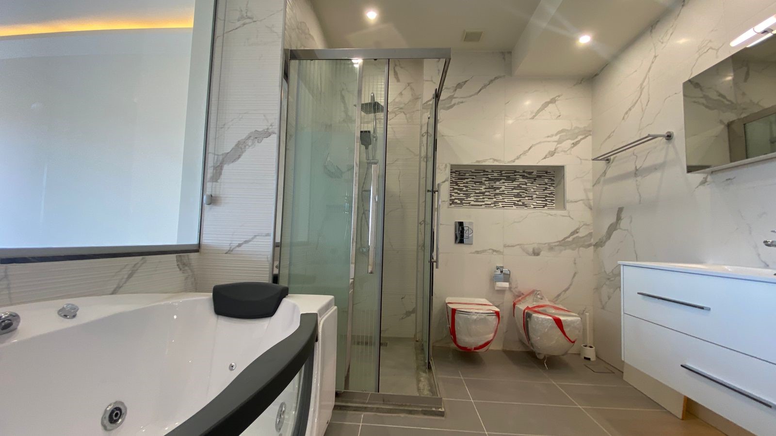 563eb5eb-3a13-4c8a-8275-5f30c480d8ed Modern bathroom with marble walls, glass shower enclosure, a large tub, and two toilets wrapped in plastic.