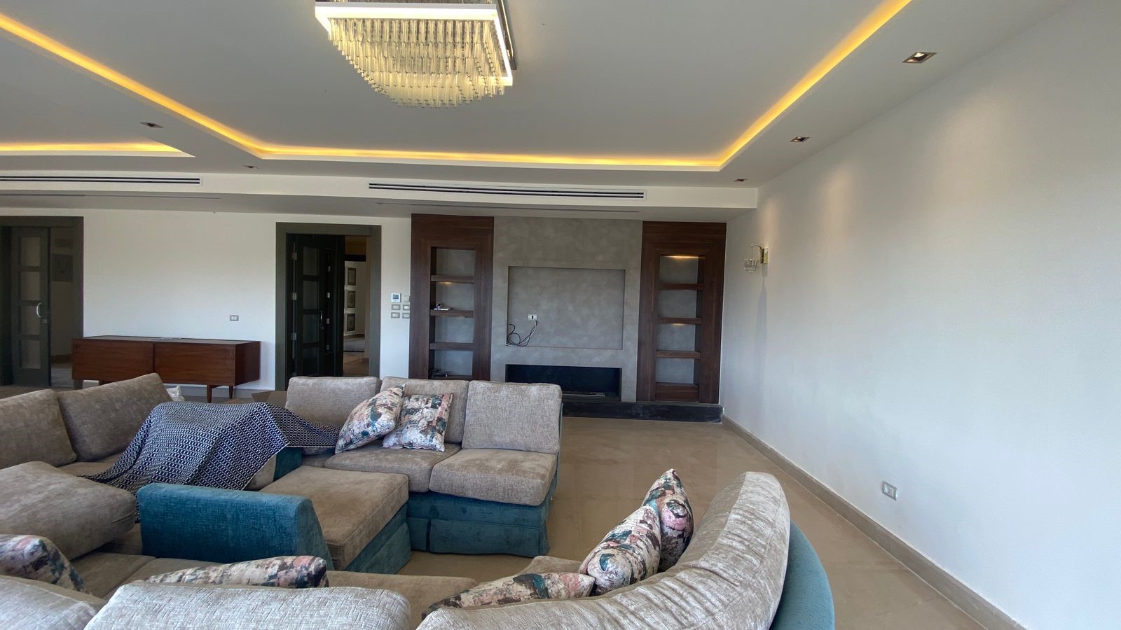 5a12e8be-64f3-47cf-a9f3-beb9d254e8fe Spacious contemporary living room featuring an L-shaped beige sofa with patterned cushions, a crystal chandelier, and a built-in fireplace wall with dark wood panels.
