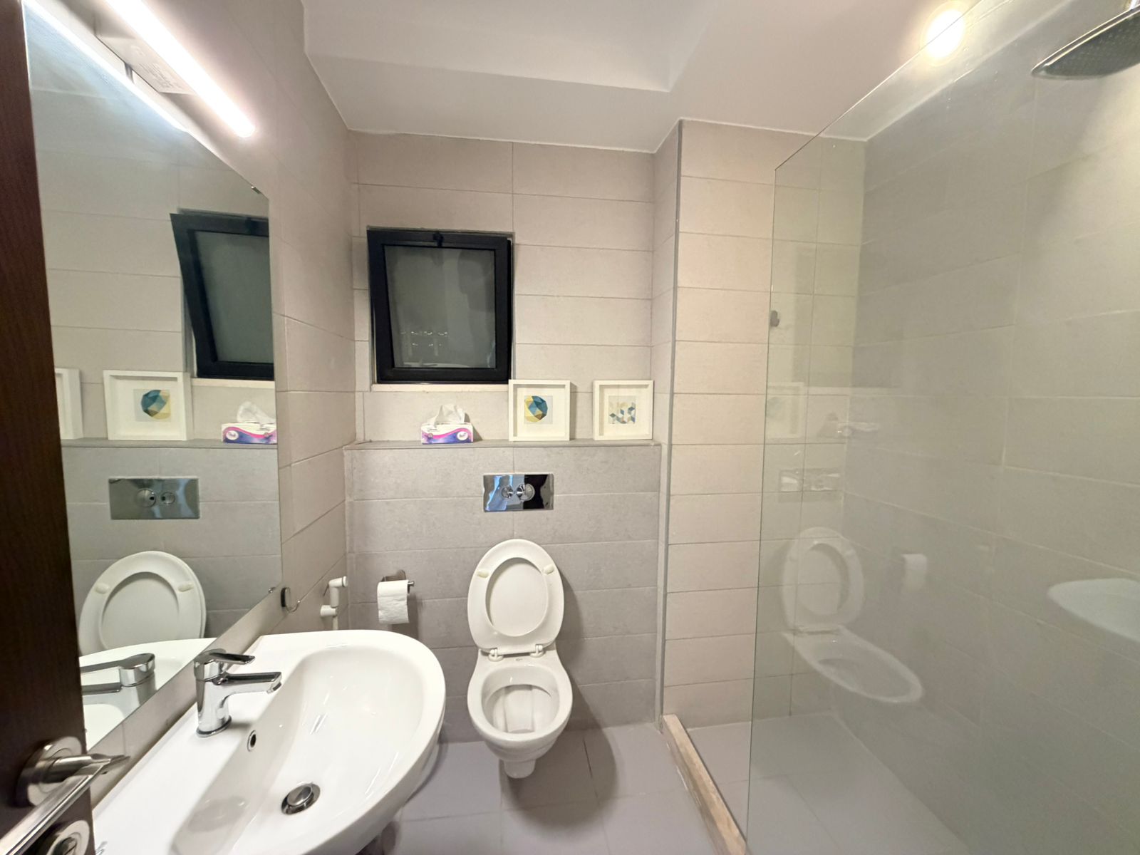 64a7e287-0d53-4a4f-a525-bb98a0406b49 Grey-tiled bathroom with a white sink, wall mirror, and an open toilet. A small window sits above a ledge holding tissue boxes, with a glass shower enclosure on the right.