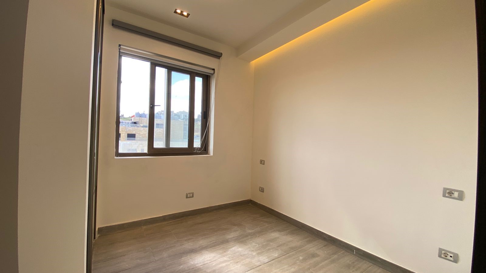6fb9a2fc-9aac-4dd3-8512-4ebdf6ca4910 Empty small room with light wood floor, white walls, and a large window showing buildings outside; built-in wardrobe on the left and a warm, recessed ceiling light strip.