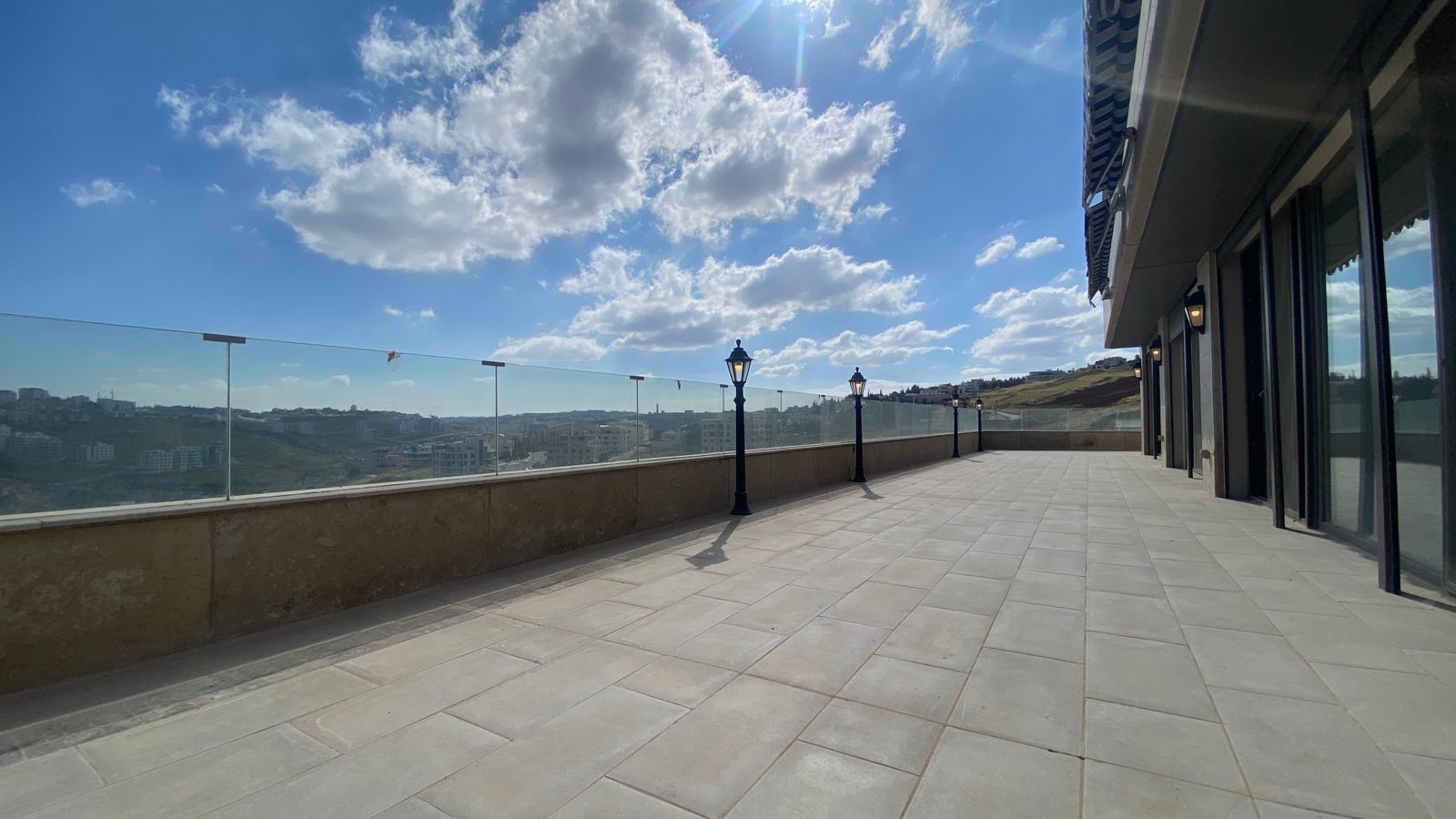 717f45db-7872-46c3-bade-6b104cf2334f Sunny rooftop terrace with a glass railing and lamp posts, overlooking a hilly cityscape and blue sky with clouds.