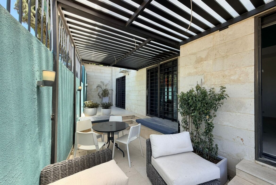 Covered outdoor patio with wicker sofas and white cushions around a round black table, beige stone walls, and potted plants.