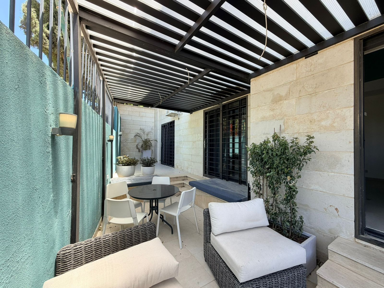 7aa8c7b1-5f54-48d3-aafa-636feb442e92 Covered outdoor patio with wicker sofas and white cushions around a round black table, beige stone walls, and potted plants.