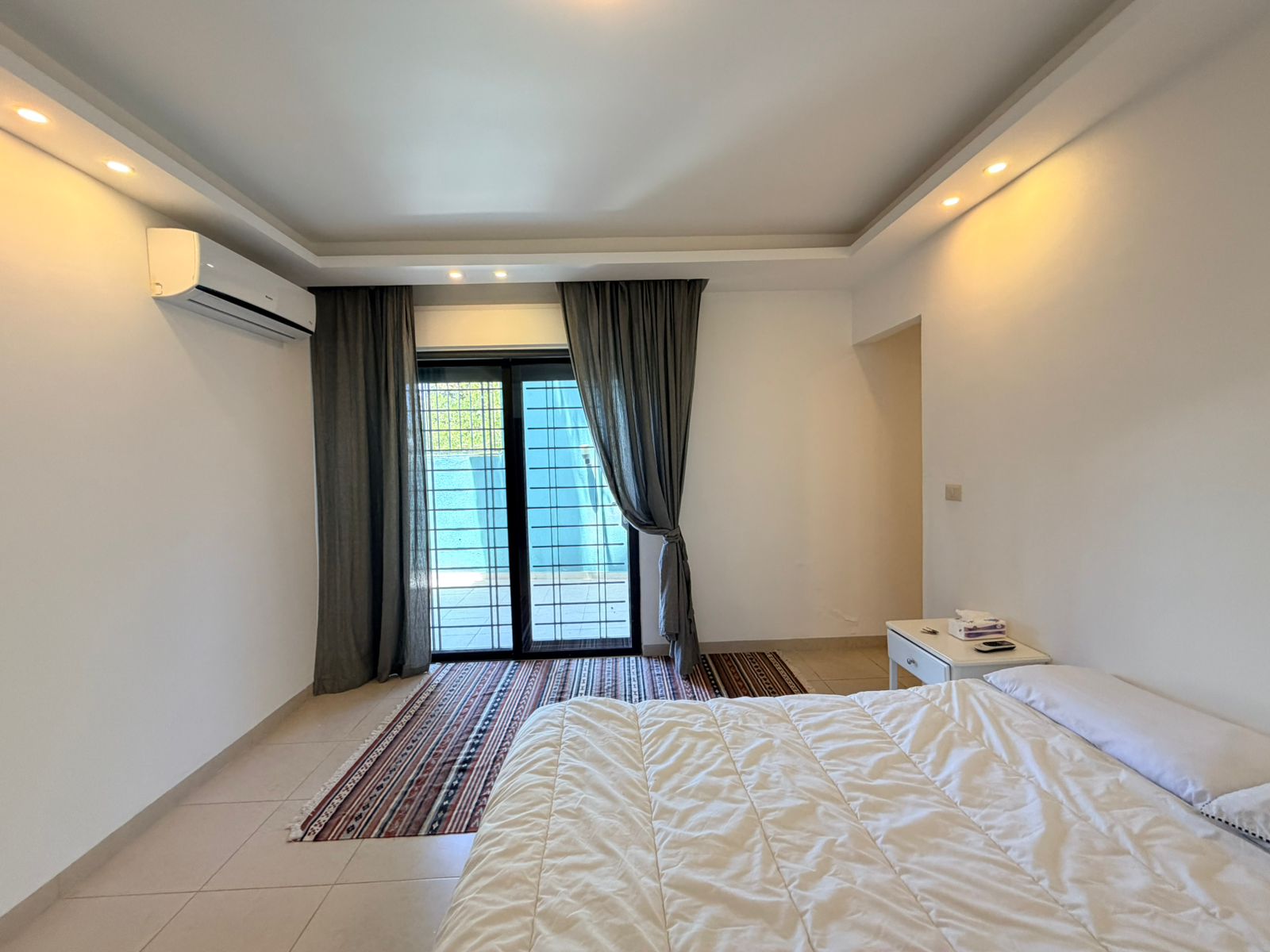 86124696-b2aa-44c2-ada1-890ee5c00eb0 Bright bedroom with white walls, a bed in the foreground, and a glass balcony door with gray curtains to the back left. An air conditioner sits on the left wall above the bed area.