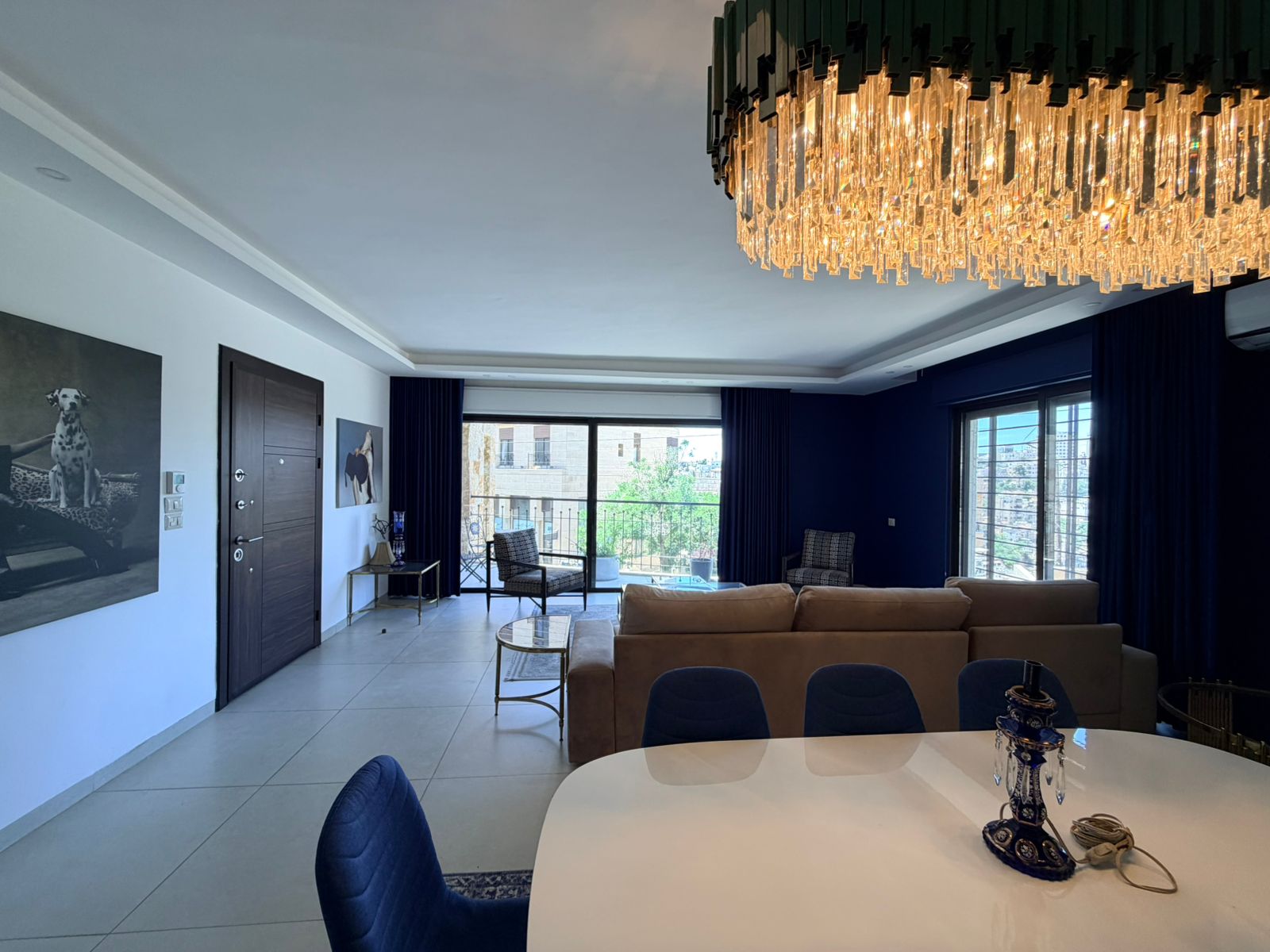 9b7a251a-017b-4f72-b497-8b0e285ae51c Modern living room featuring a large crystal chandelier, blue accent wall, beige sofa, and a round white dining table in the foreground. A balcony door and city view are visible outside.