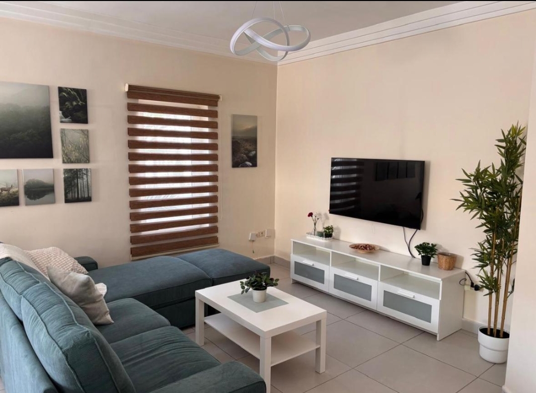 WhatsApp Image 2026-04-25 at 124140 PM Living room with a teal sectional sofa, white coffee table, wall-mounted flat-screen TV, and a white media console near a tall plant.