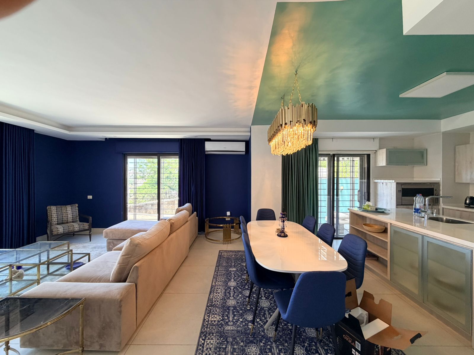 bc8195b7-7f26-44a4-b124-9eb27a5b357c Open-concept living and dining room with navy walls, beige sectional sofa, glass coffee tables, and a white dining table surrounded by blue chairs under a gold chandelier. A green ceiling features art-like color blocks and sliding doors illuminate the space.