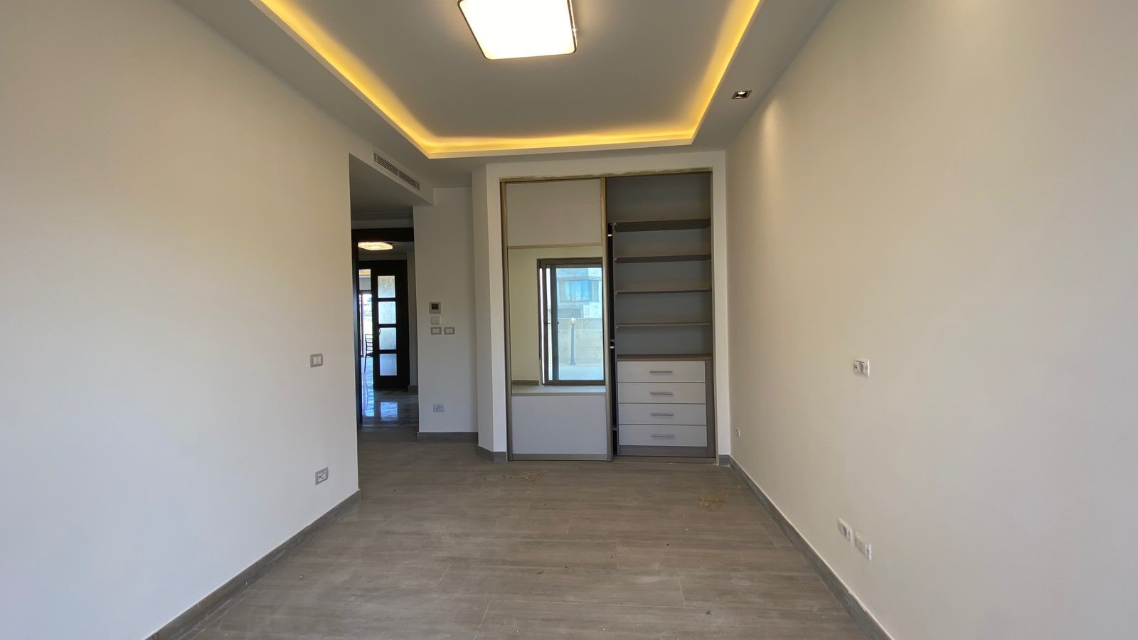 d8889e05-1ecb-4ba9-adb1-83e1ccbed4af Empty modern room with a built-in wardrobe featuring open shelves and drawers against the back wall, large window, and recessed ceiling lighting.