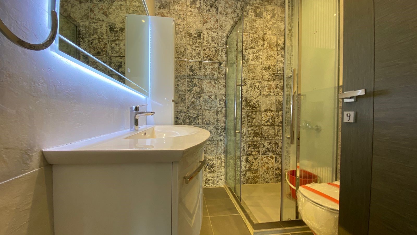 eaa78eb7-c5b8-44c5-84a3-bf372f095512 Modern bathroom with white vanity and sink, backlit mirror, patterned tile wall, and glass shower enclosure.