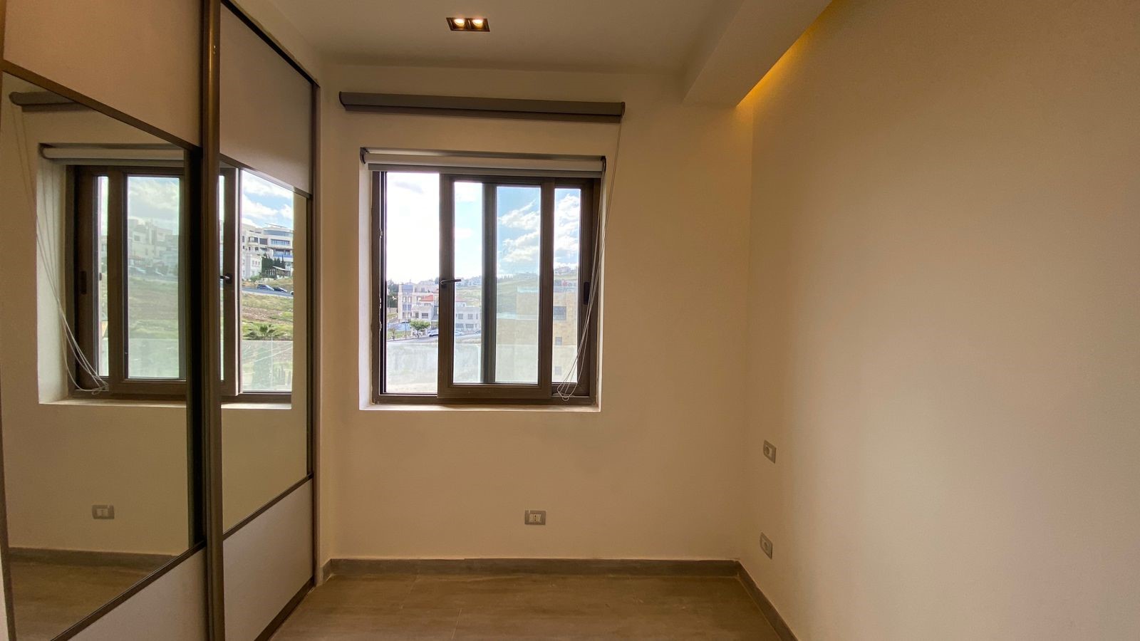 eb2dffaa-3150-409d-8be7-b40c5845dbe6 Empty modern bedroom with a large window and built-in mirrored wardrobe on the left, neutral walls, and gray tile floor.