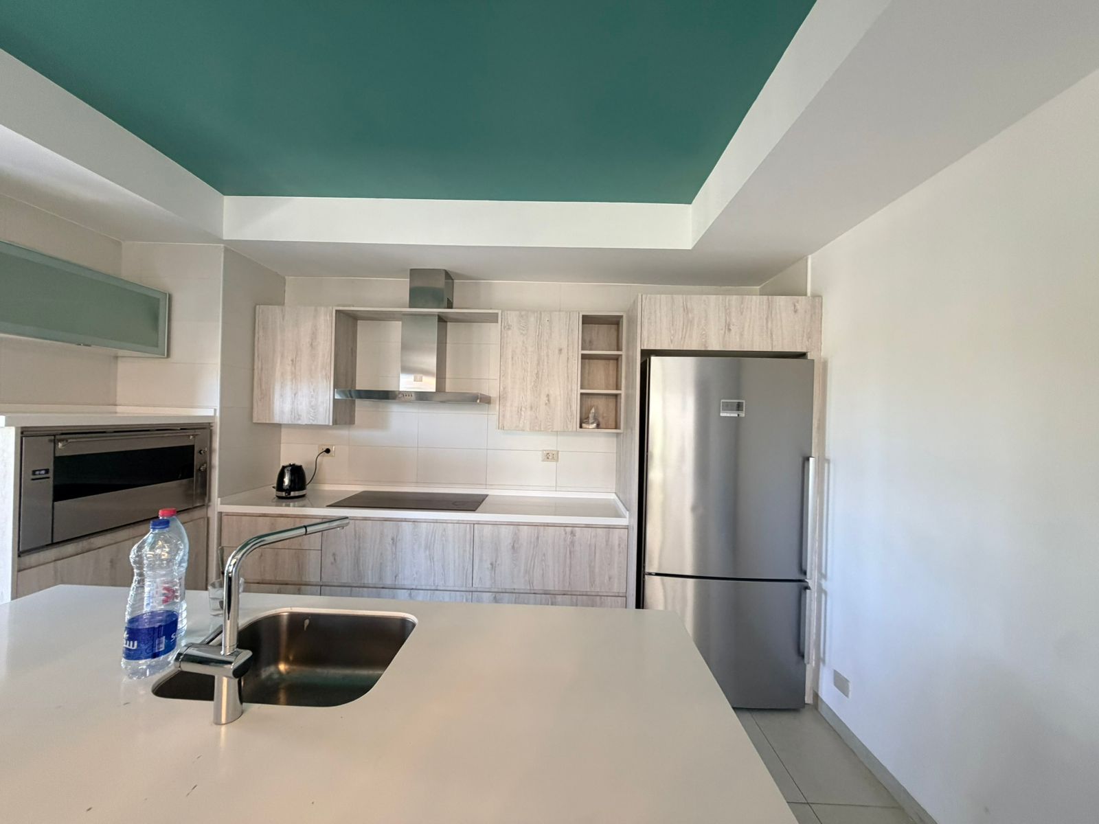 ebf315e2-2fa1-4468-a2e7-b00a56301bfb Contemporary kitchen with light wood cabinets, stainless fridge, and a teal ceiling; island sink in foreground.