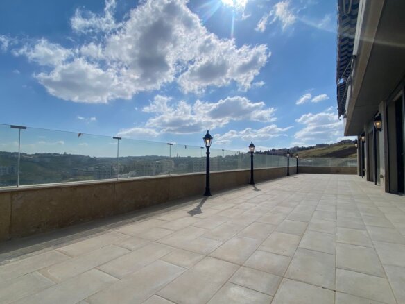 A wide outdoor terrace with a glass railing, lampposts, and a modern building on the right under a bright blue sky with fluffy clouds.