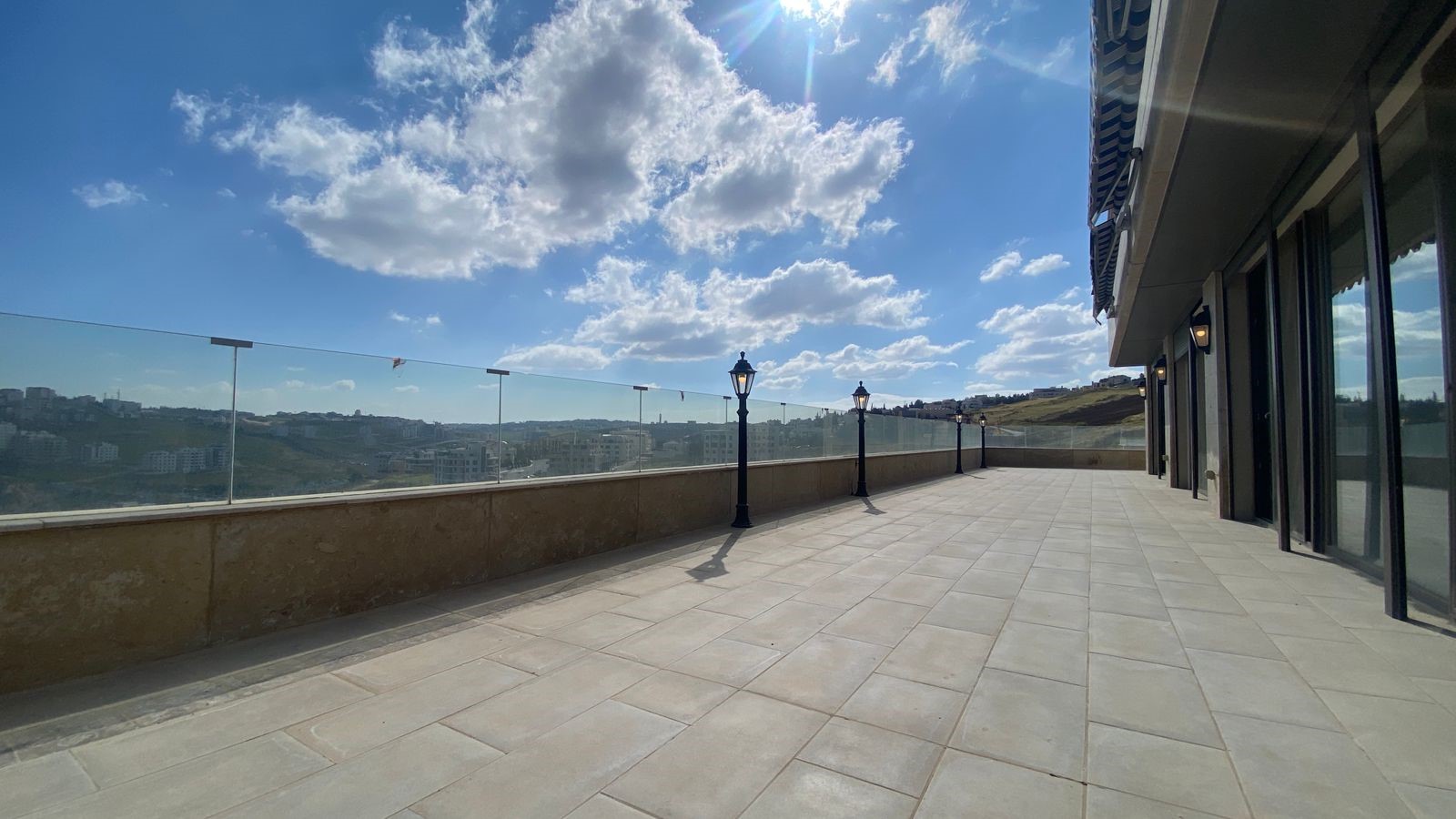 A wide outdoor terrace with a glass railing, lampposts, and a modern building on the right under a bright blue sky with fluffy clouds.
