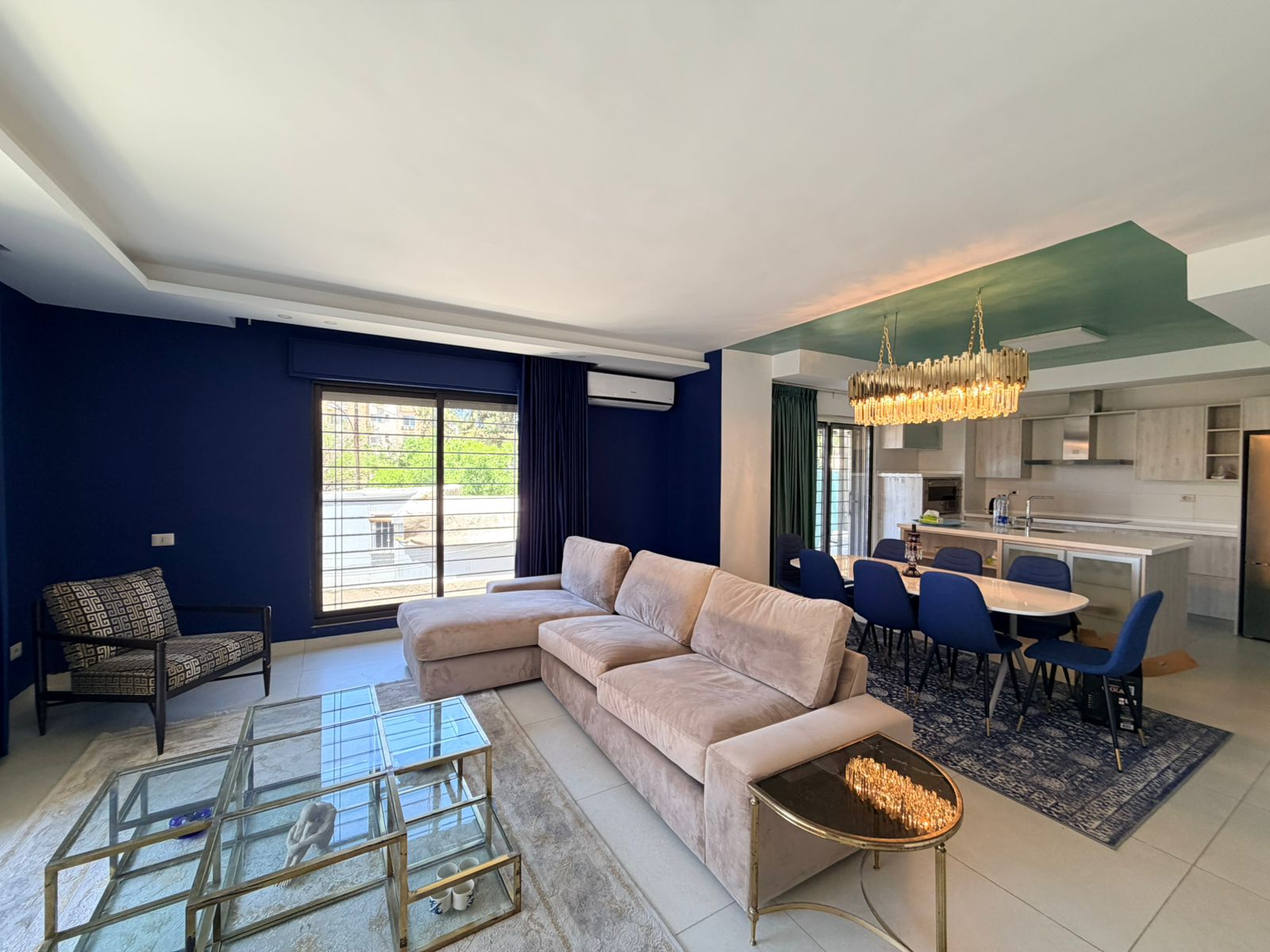 f5fbc7d4-11d7-421b-ae4e-e46215e20ea0 Open-plan living room and kitchen: navy blue wall, beige sectional sofa, glass coffee tables, blue dining chairs, crystal chandelier, and a green ceiling accent with large windows.