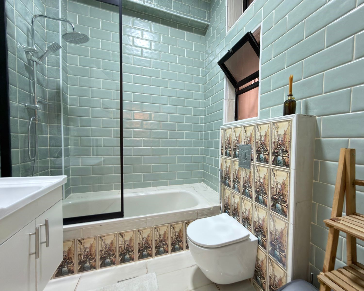 15527632-7fdb-4cee-8a6a-e3c8bb05f69d Bathroom with teal subway tiles, glass shower door, and built-in tub; decorative tiled cabinet beside a wall-hung toilet.