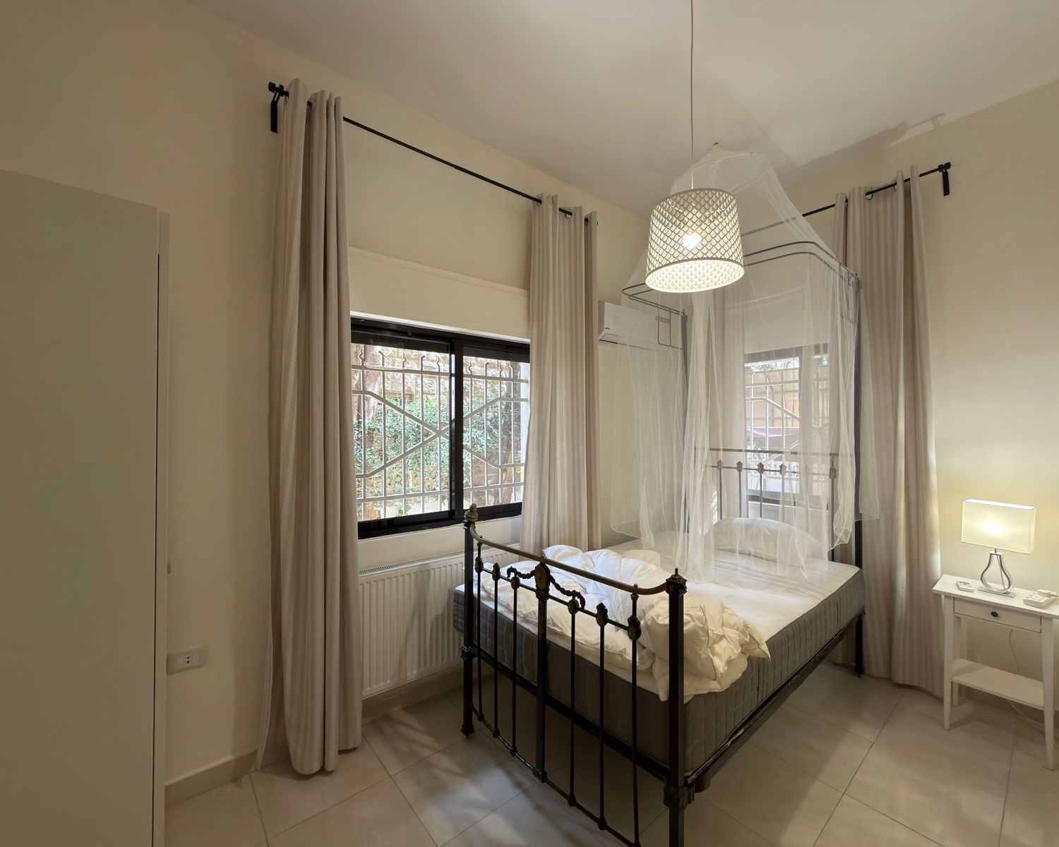 201254ae-c32f-45a3-ac7f-5772f1498166 Bright bedroom featuring a black metal four-poster bed with a sheer canopy and white bedding, opposite a window with light curtains.