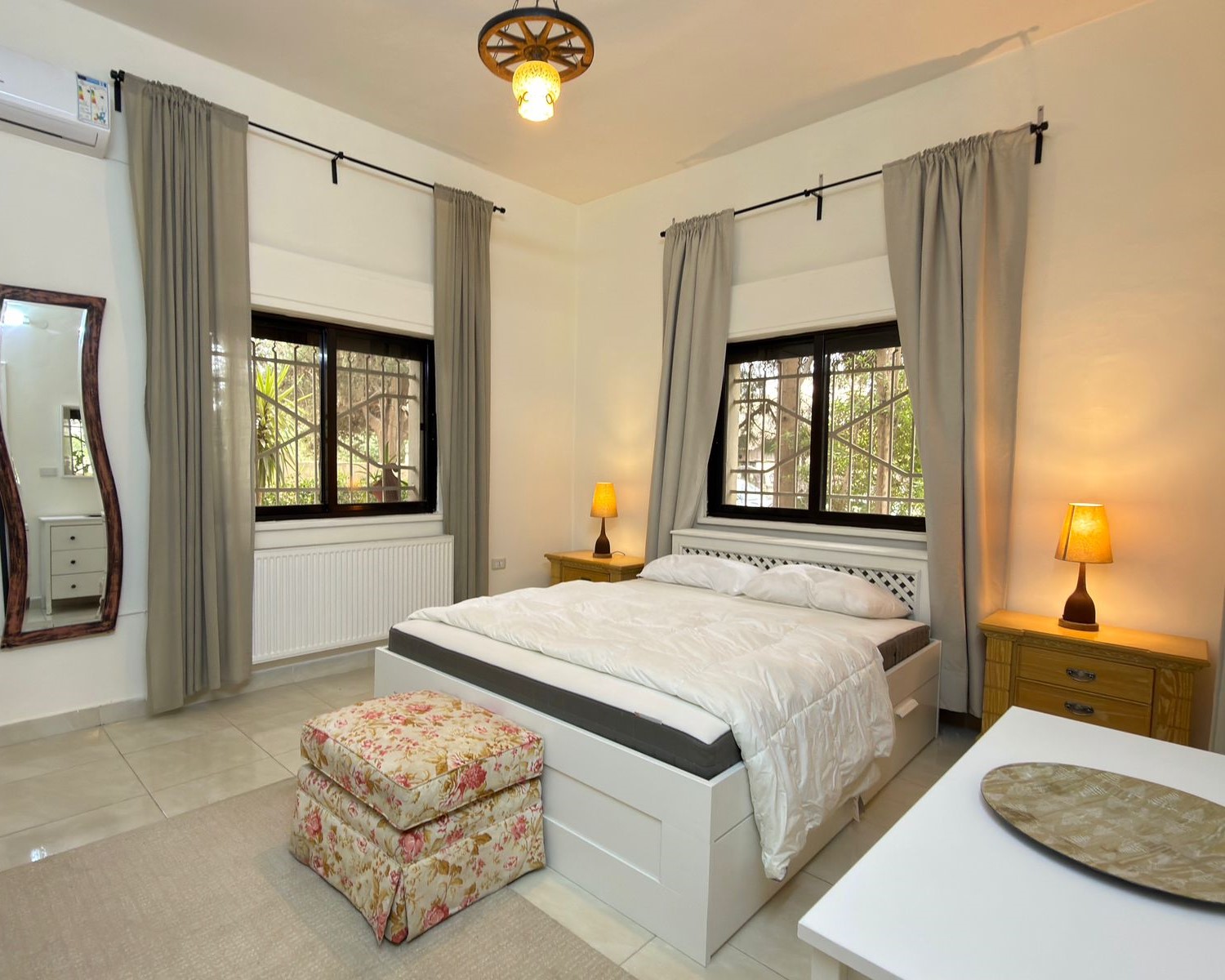 2b1e46c1-7f7e-4b18-8961-b2033bbf8ddb Bright bedroom with white bed, two wooden nightstands, and warm lamps on each side; gray curtains by two windows.