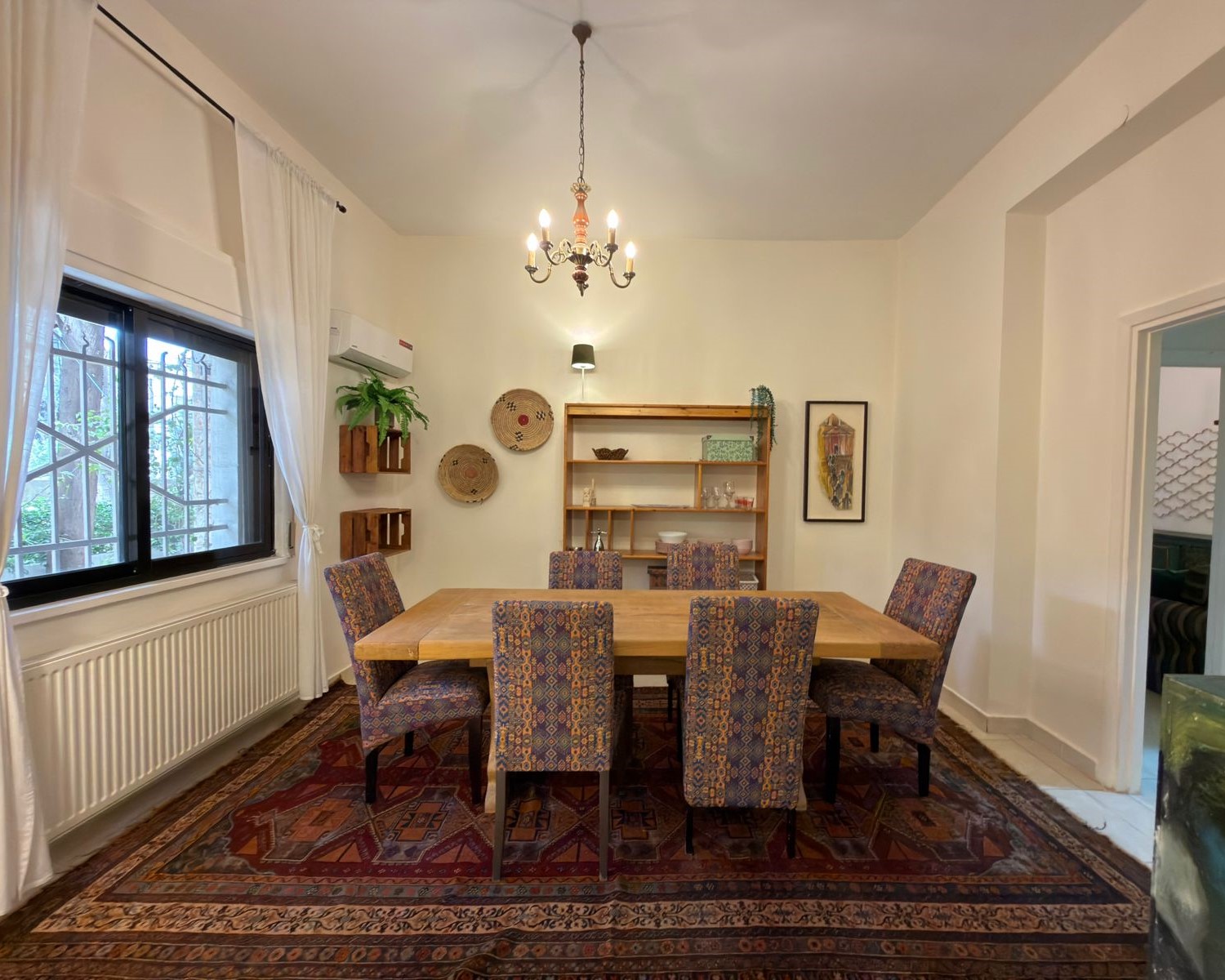 2dfdc9d1-dab7-47b1-99de-c6ebeca73e56 Bright dining room with a large wooden table and six patterned upholstered chairs on a decorative rug under a chandelier.