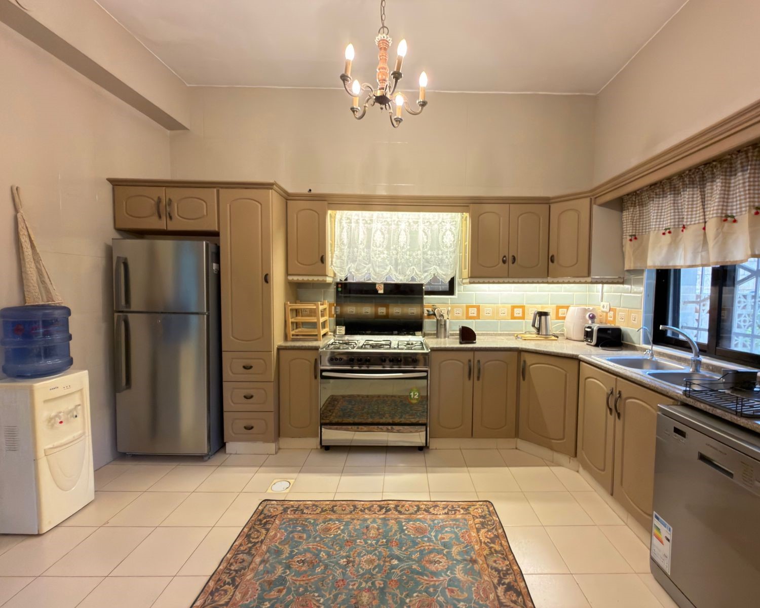 3329e977-08b1-4218-8ea7-24e00eb605ce Beige U‑shaped kitchen with stainless steel fridge, gas range, and dishwasher; tiled floor and a decorative rug in front of cabinets.