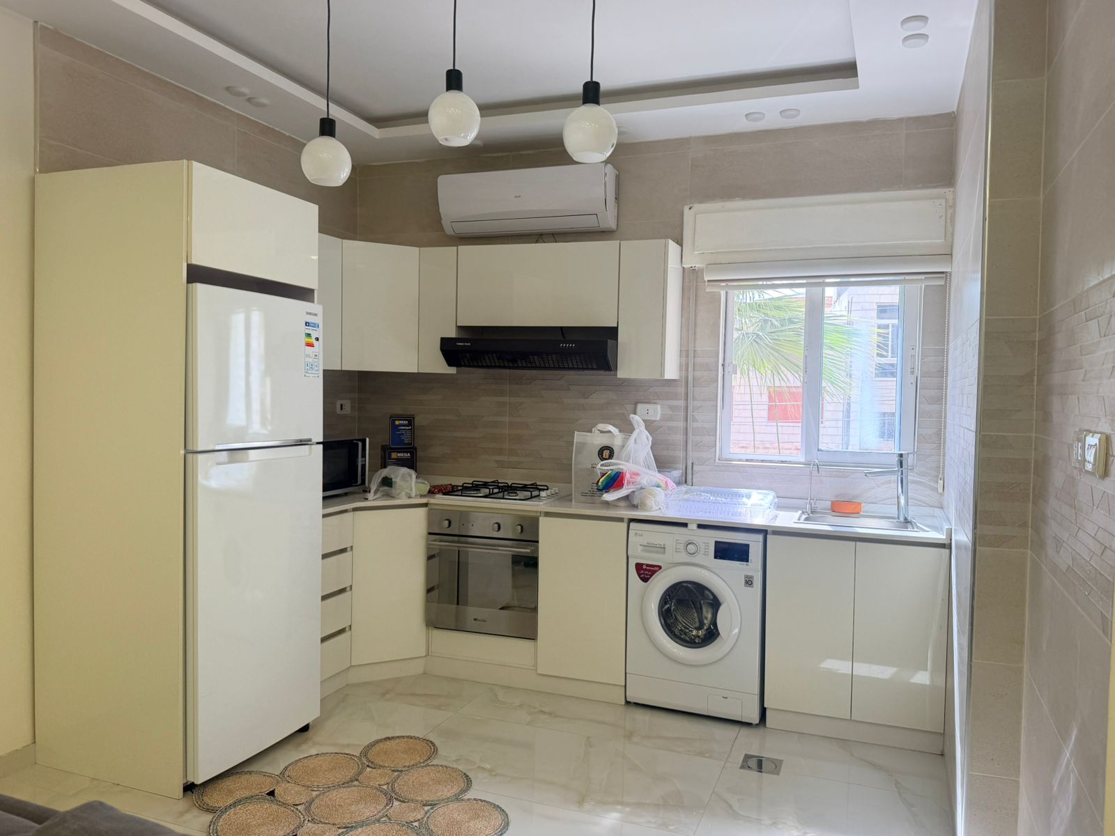 3ba78fc3-8695-435d-93cd-32a597bdffe4 Modern white kitchen with tall fridge, built-in oven, gas cooktop, range hood, and washing machine under counter next to a window.