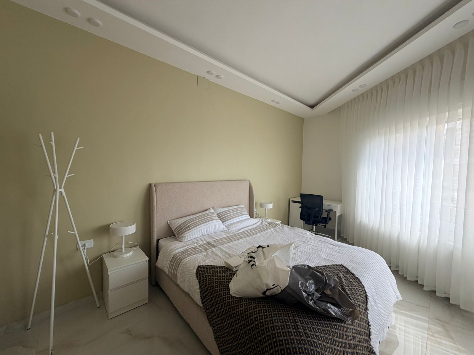 3e6611f1-79bb-46ca-81b5-30dcafe5a6a8 Bright bedroom with beige upholstered bed, two white nightstands, and lamps; coat rack on the left and a desk with chair by a large window with sheer white curtains in the background.
