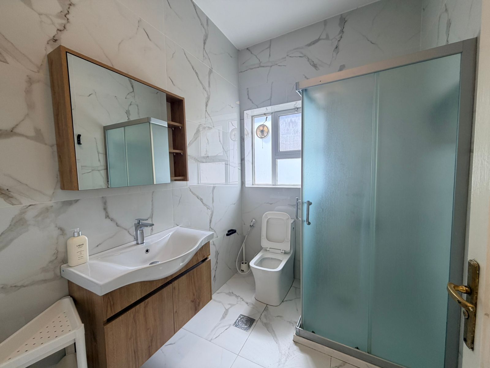 3eb03236-ca0f-48db-a8c0-ebd5ed383b75 Bathroom with marble walls, wooden vanity and sink, wall mirror cabinet, toilet, and a frosted glass shower door.