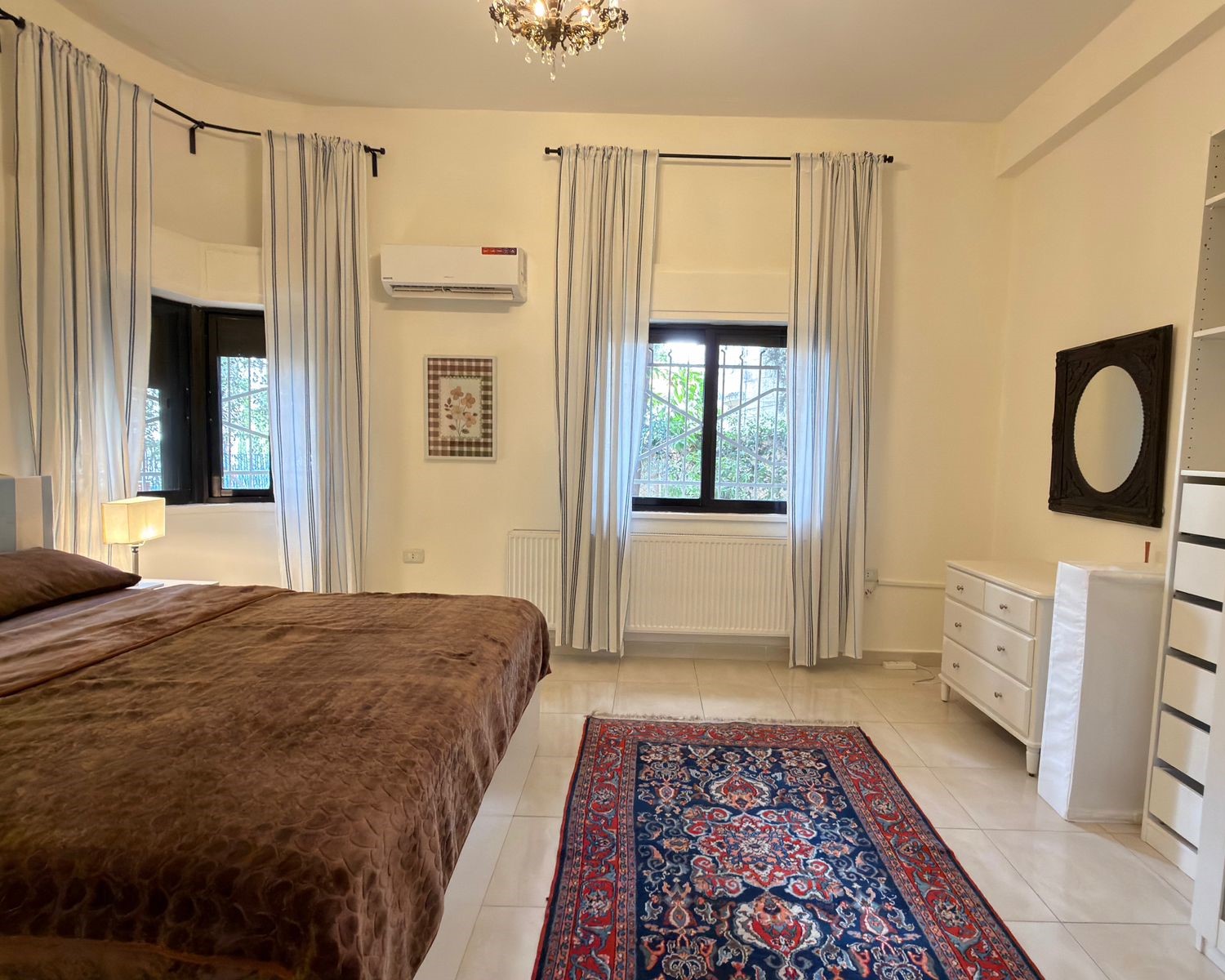 554e2354-d72a-451b-b783-82e458e8d29d -bedroom with a large brown quilted bed, white nightstand, and lamp on the left; blue striped curtains and a chandelier above