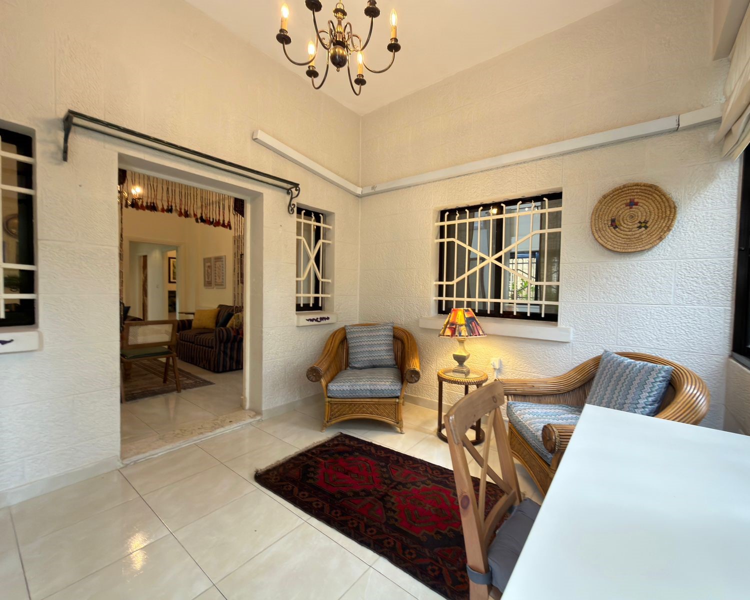 686095cf-61cf-44bb-bd62-8bf4f4fa07c7 Bright sunroom or enclosed porch with wicker seating, a small table lamp, and a patterned rug on tiled floor.