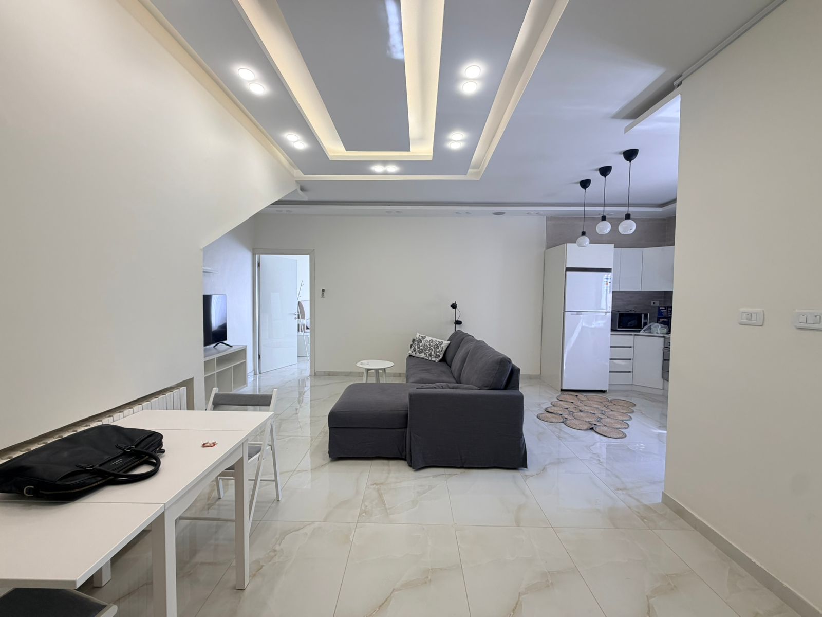 74bd568c-e044-40df-a9a2-5c7b447e6448 Open-concept living area with a gray L-shaped sofa facing a TV on a white console, white dining table and chairs, and a modern kitchen in the background.