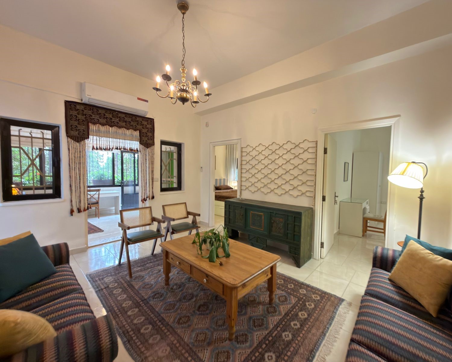 8add8915-35f9-45b9-82c2-4504433dbde6 Living room with striped couches, wooden coffee table, ornate rug, chandelier, and a dark green cabinet against the wall.