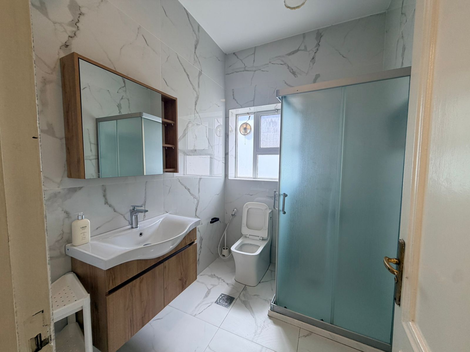 a18bc287-6b16-41ee-97cd-70a8458a35f6 Bathroom with marble walls and floor, wooden vanity with integrated sink, wall mirror cabinet, toilet, and a frosted glass shower enclosure.