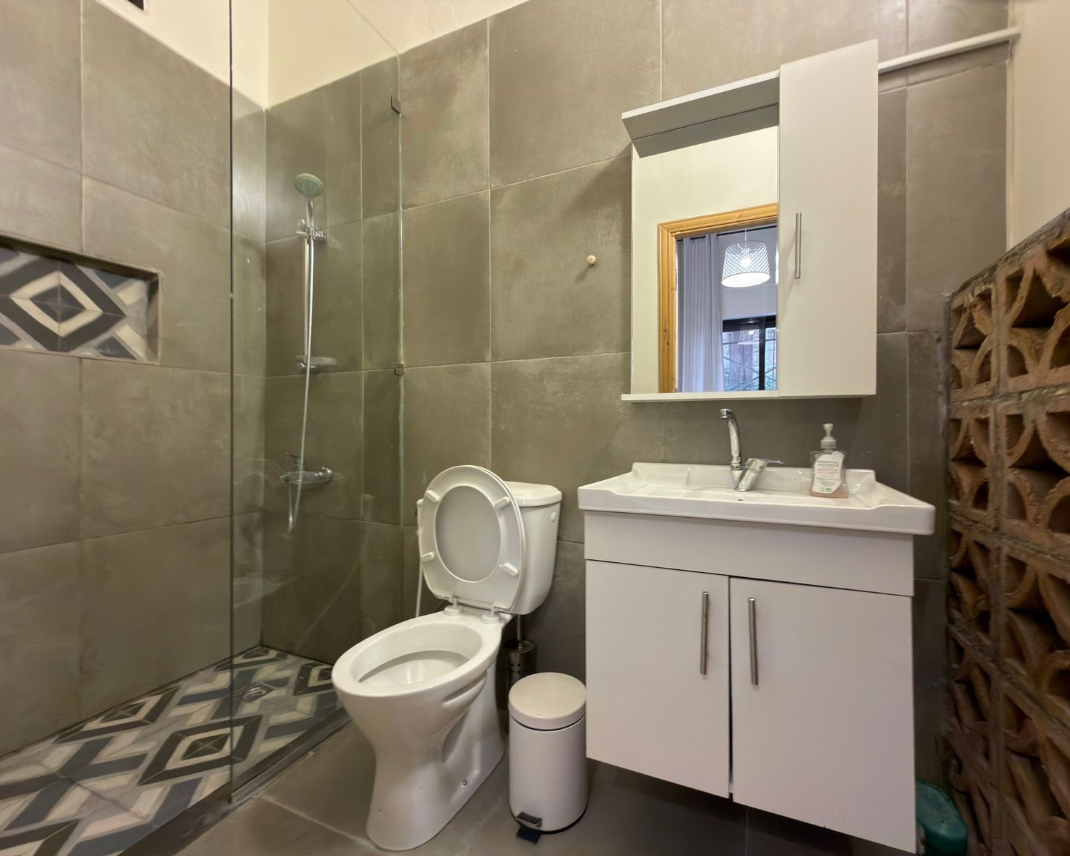 a1ff3a69-4cac-48aa-a238-e9bdfe67bd34 Modern bathroom with glass-enclosed shower, toilet, and white vanity mirror cabinet against gray tiles.