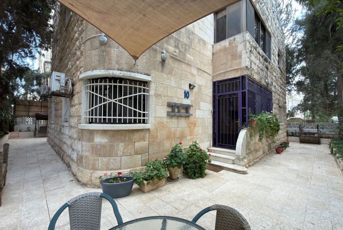 Stone building with a gated rounded window, purple front gate, and potted plants in a sunny courtyard. A glass table and chairs are in the foreground under a shade canopy.