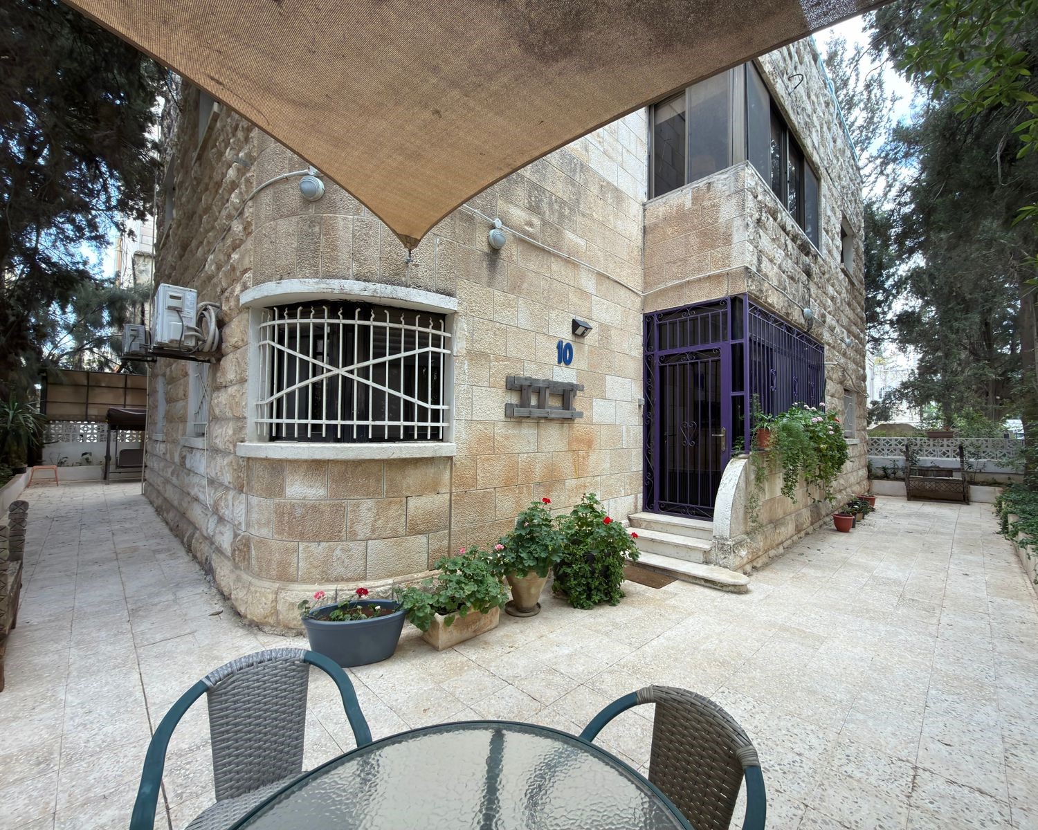a3eec766-f05e-4029-92c3-8a4d59f0662a Stone building with a gated rounded window, purple front gate, and potted plants in a sunny courtyard. A glass table and chairs are in the foreground under a shade canopy.