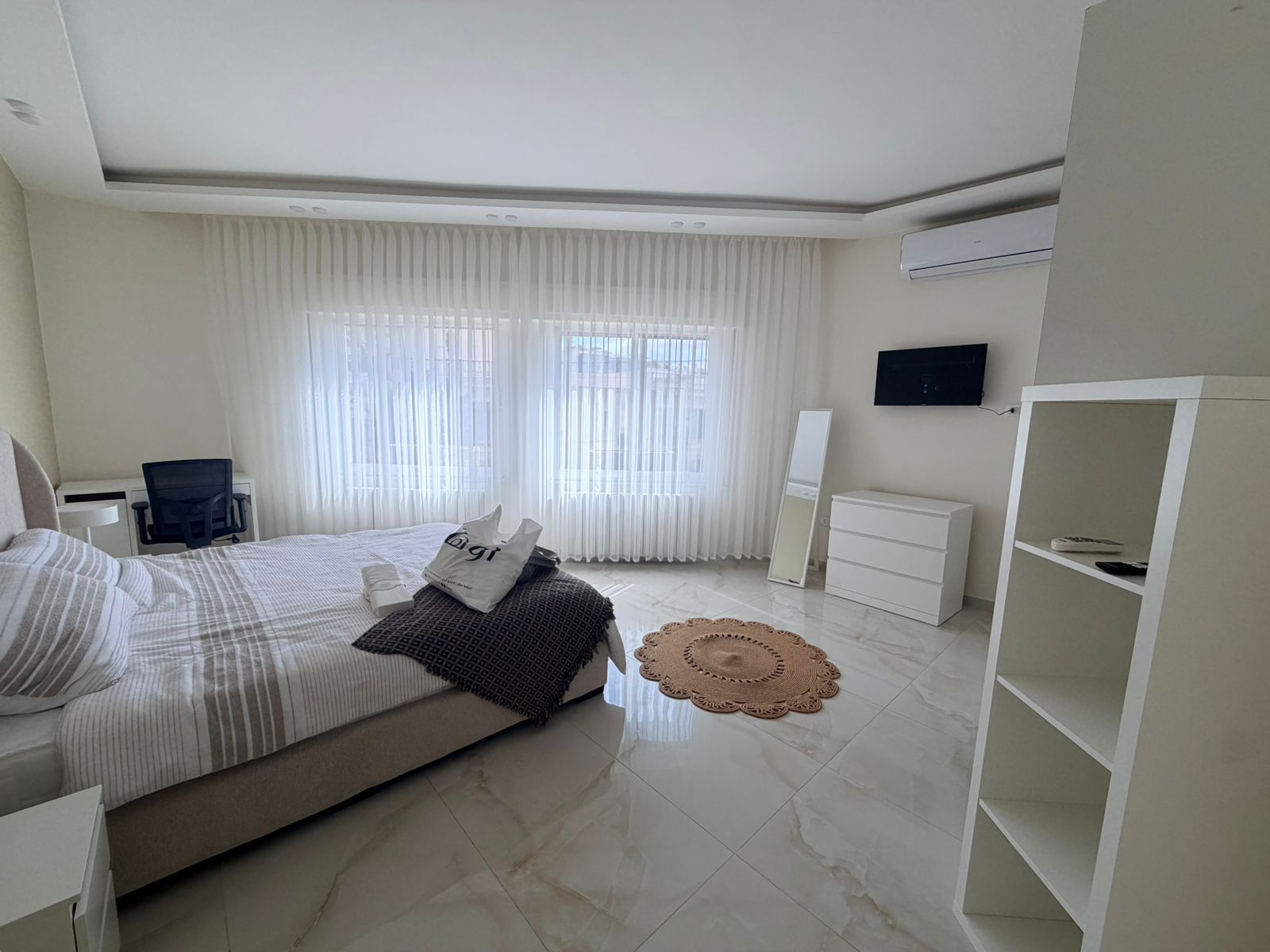 f72c446c-f92e-41fe-b882-74d57c9320db Bright modern bedroom with a large window and sheer white curtains, a bed on the left, and white furniture across the room.