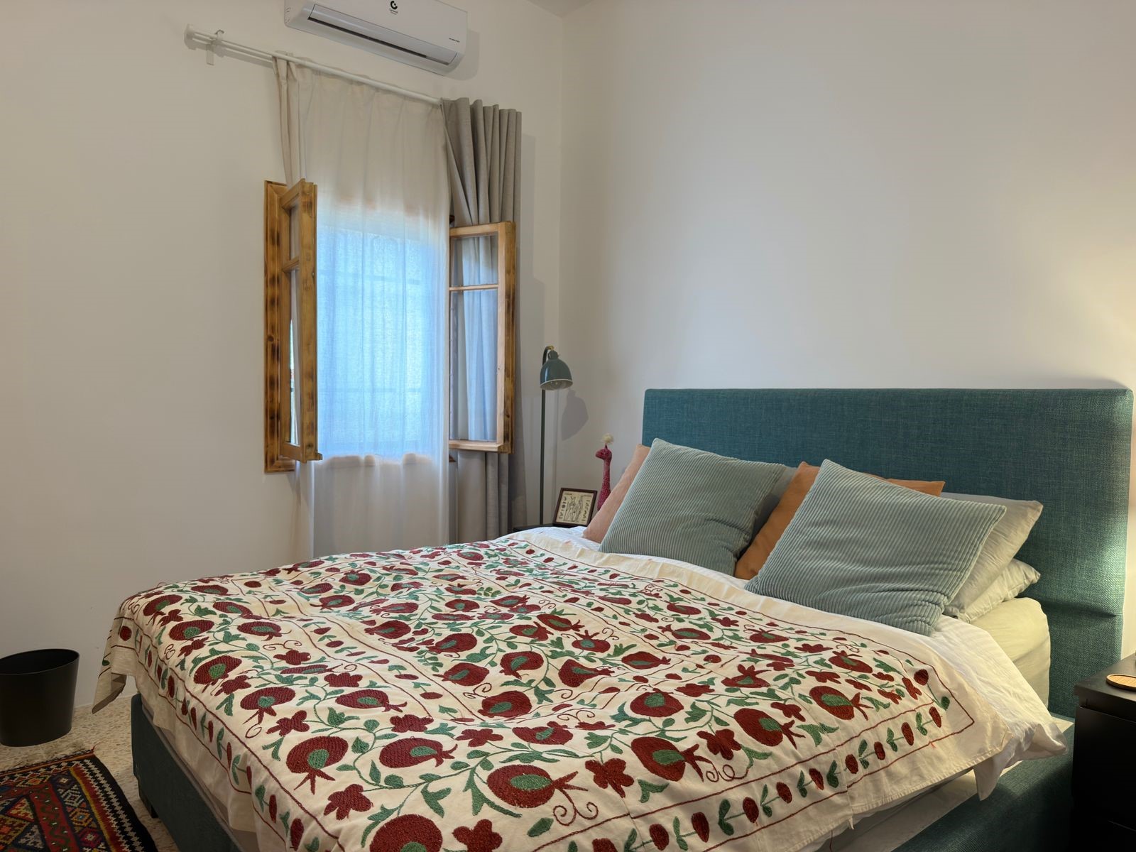 0020062f-bb9a-4420-b7a9-ab12ea490394 A neatly made bedroom with a teal upholstered headboard, floral quilt, and multiple pillows; window with wooden shutters and sheer curtains beside a standing lamp and small side table.