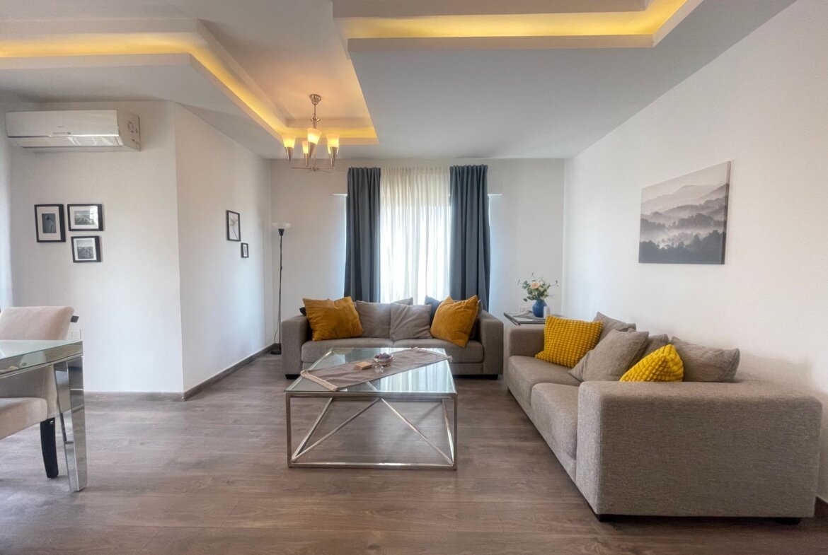 Modern living room with gray sofas, yellow cushions, and a glass coffee table under warm ceiling lights.