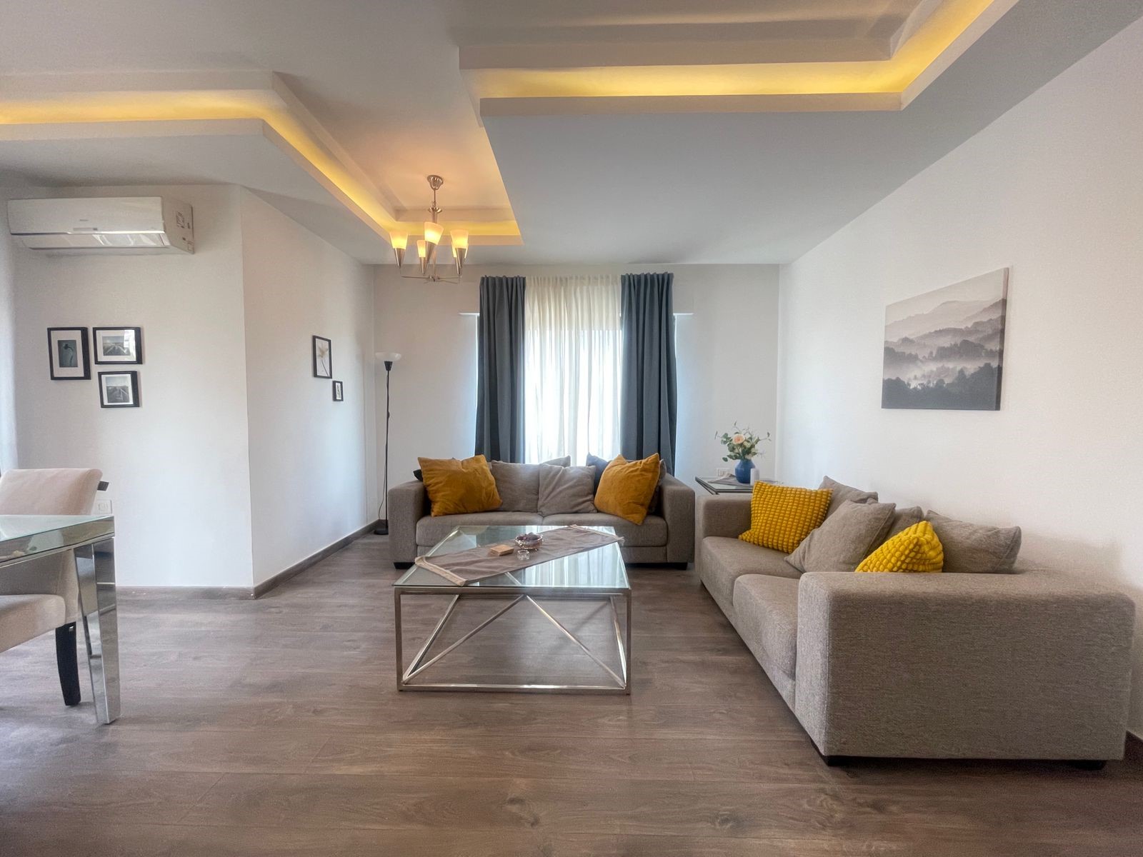 0056e92a-f748-4a72-8f30-5d88c1047b14 Modern living room with gray sofas, yellow cushions, and a glass coffee table under warm ceiling lights.