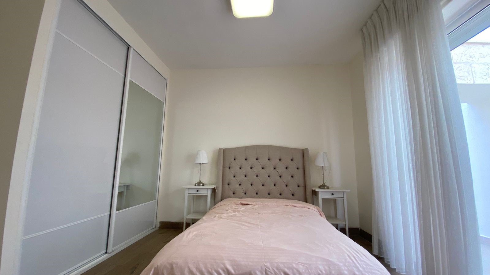 00b483f5-acc0-49ba-92bf-e416b37dfa05 – Copy Bedroom with a single bed, pink duvet, tufted beige headboard, two white nightstands with lamps, and a large sliding-door wardrobe.