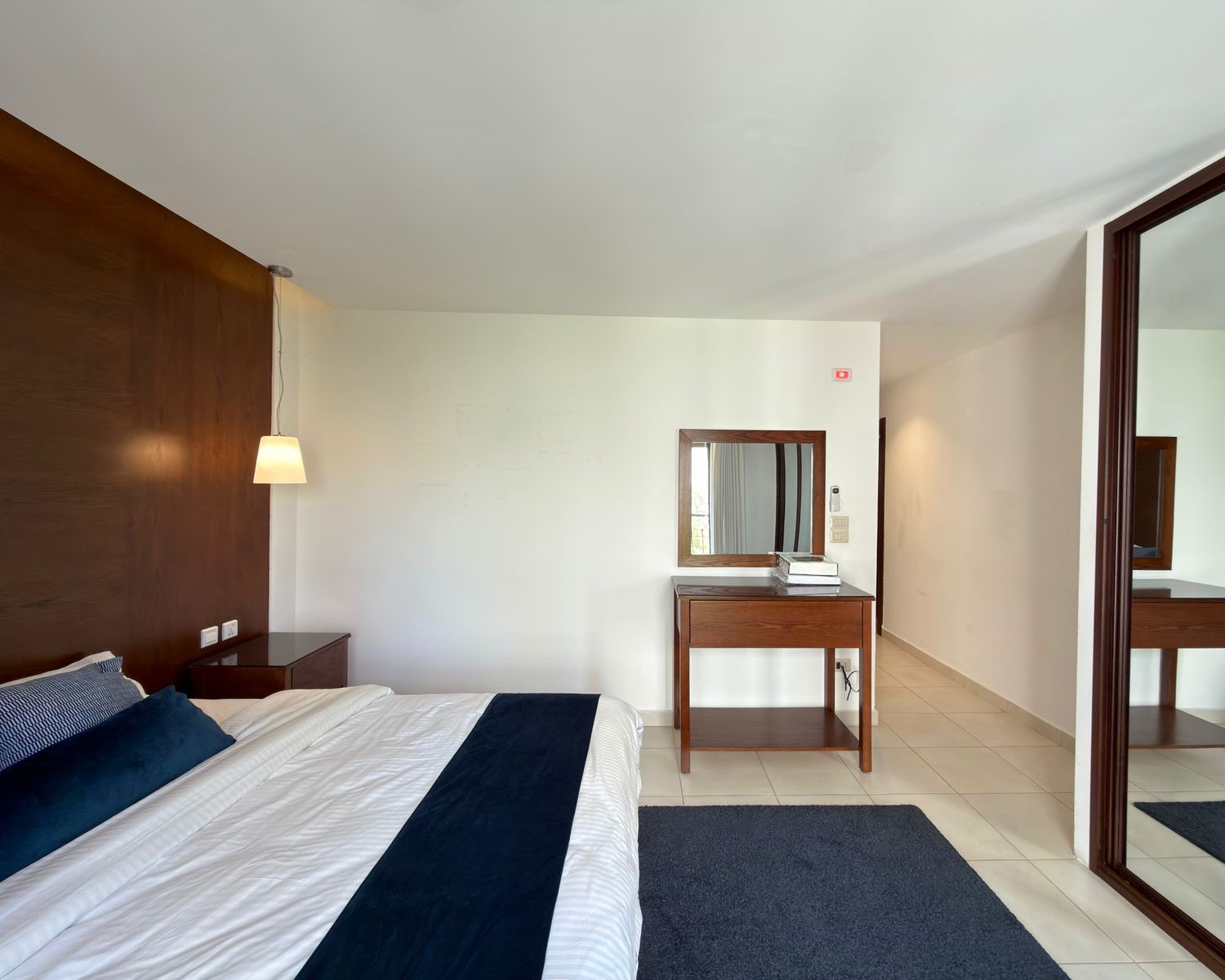 01c970a6-a122-4faf-9a70-b7eaa96e1dd4 Hotel bedroom with white linens, navy runner, and dark wood headboard against a white wall.