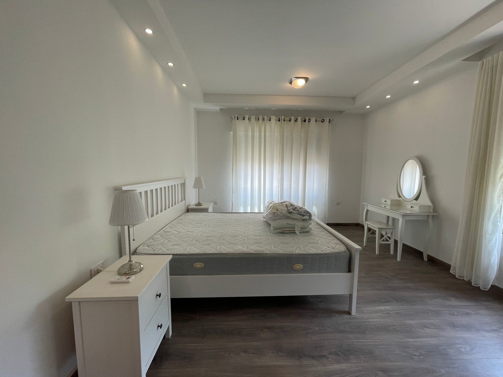 01d6eeb3-c9b1-4ddf-b44a-71444e753b2a Bright white bedroom with a queen bed, two white nightstands, and two lamps; sheer curtains at the window.