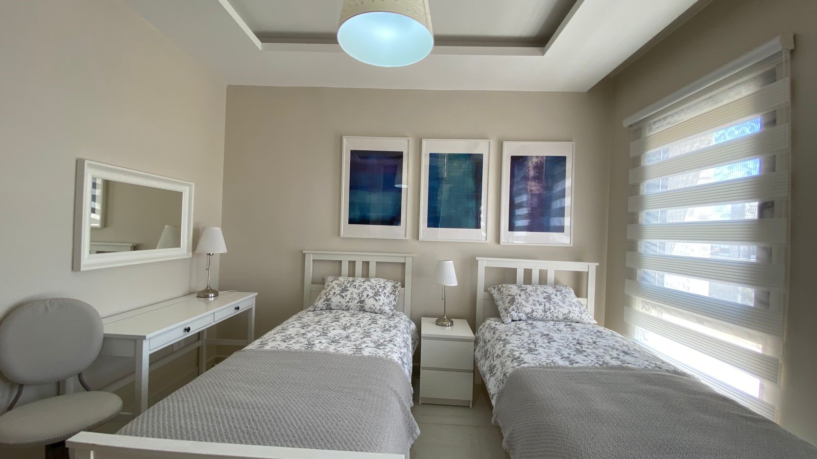 02dd14f9-bcf6-486b-9325-5844b364f057 Twin bedroom with two white-framed single beds, matching nightstand and lamps, beige walls, and three blue abstract prints above the bed wall art above.