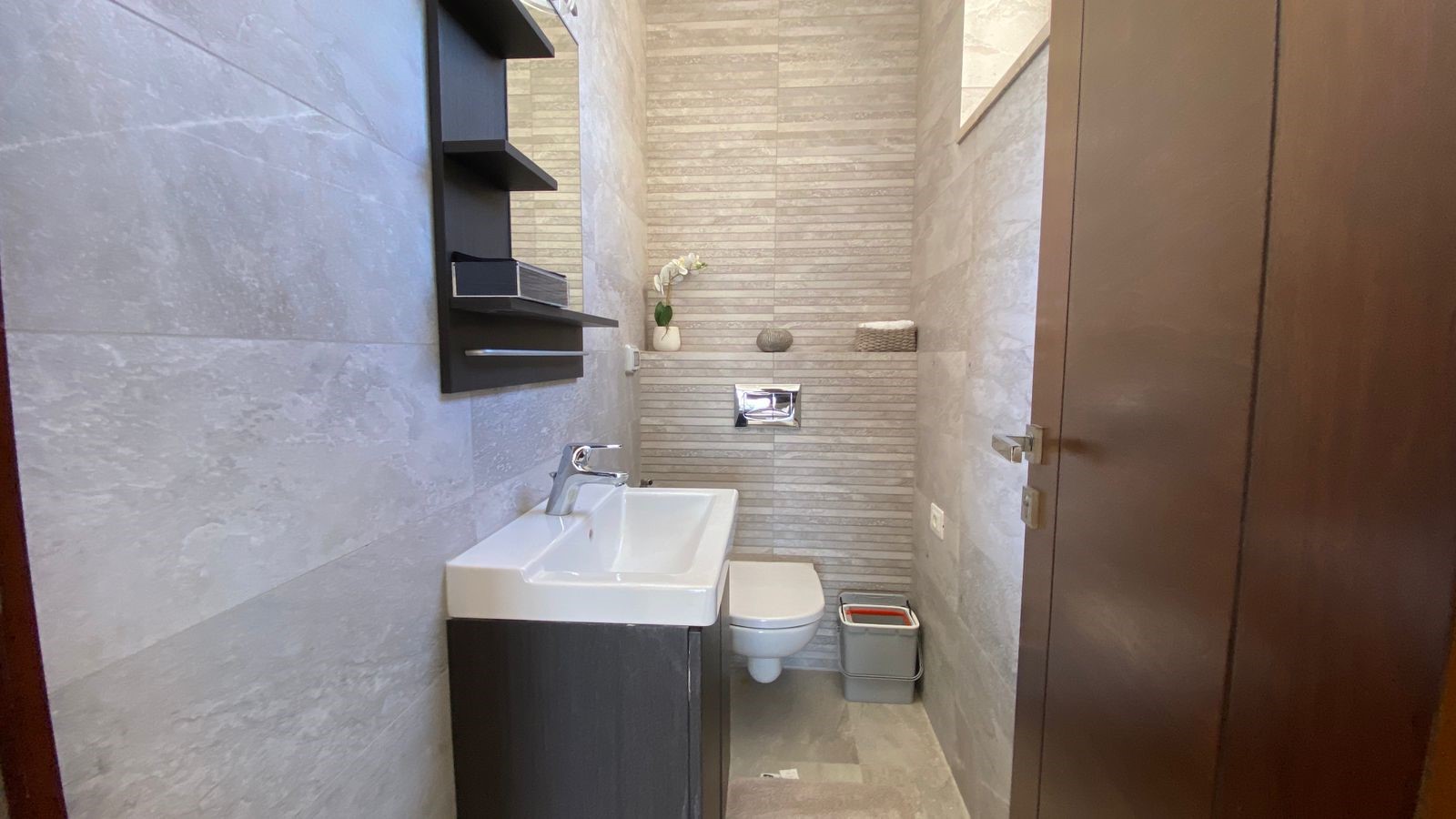 042f06d1-8d78-4855-acee-22c2d2e99620 Narrow modern bathroom with a white rectangular sink, dark vanity, and wall-mounted toilet against gray tiled walls