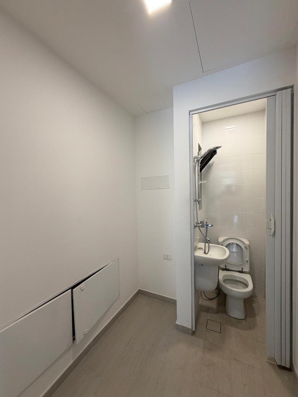 05cb3d6a-23ad-493a-976d-11069f99a4bd Small utility bathroom seen through an open door: toilet with lid up and a compact wall-mounted sink, plus a handheld shower on the tiled wall behind.