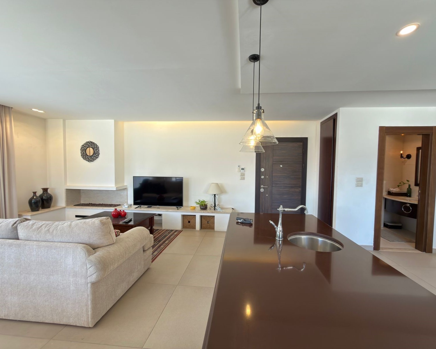 064eb1b9-a277-4259-a064-98a237dfd4db Open-concept living room with a beige sofa facing a flat-screen TV and a white entertainment unit beside a doorway to the kitchen island area.
