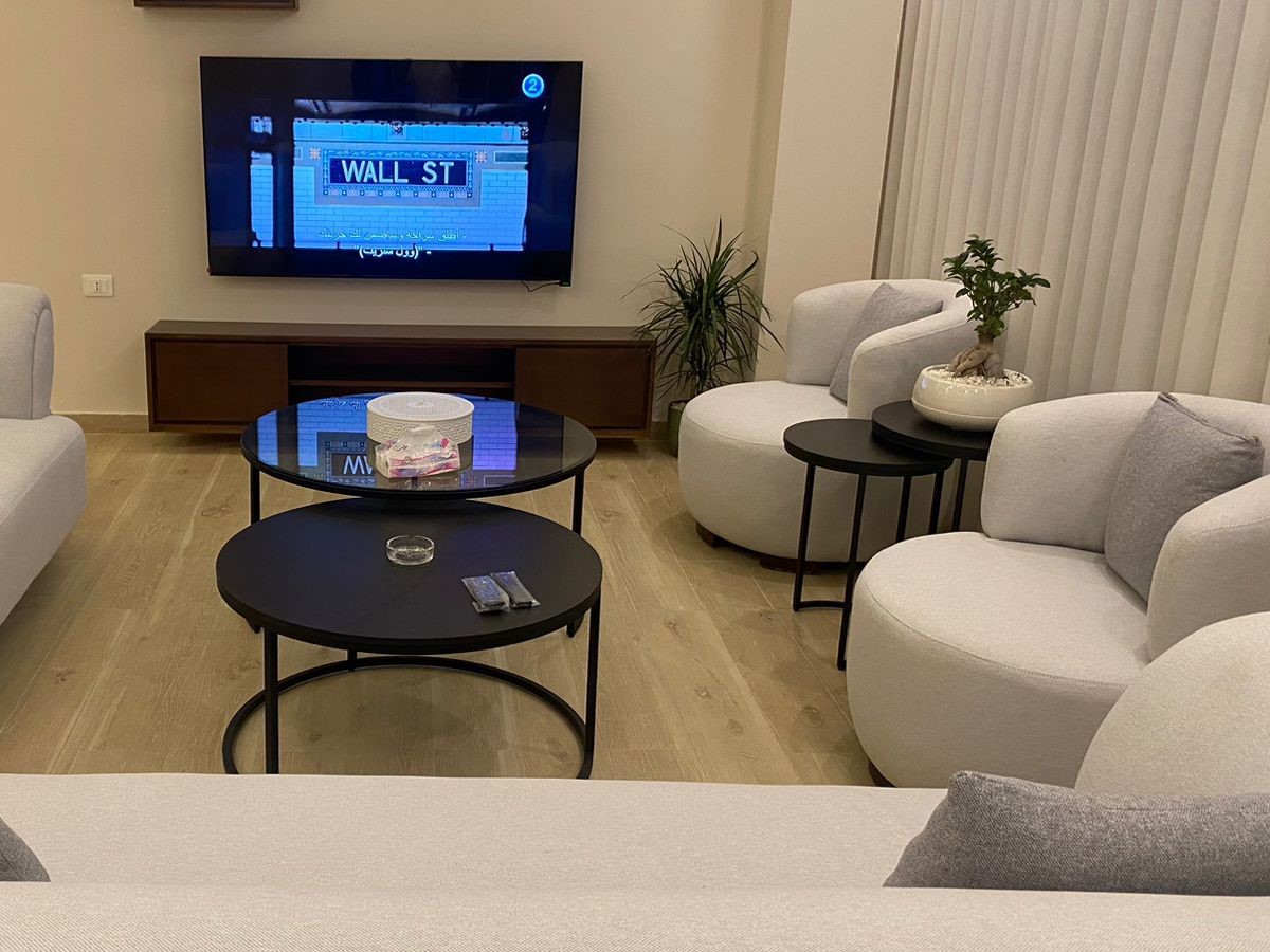 06a45267-838a-4ae0-9067-deac81f018a2 Living room with a wall-mounted TV showing Wall St, round black coffee tables, and cream curved sofas.