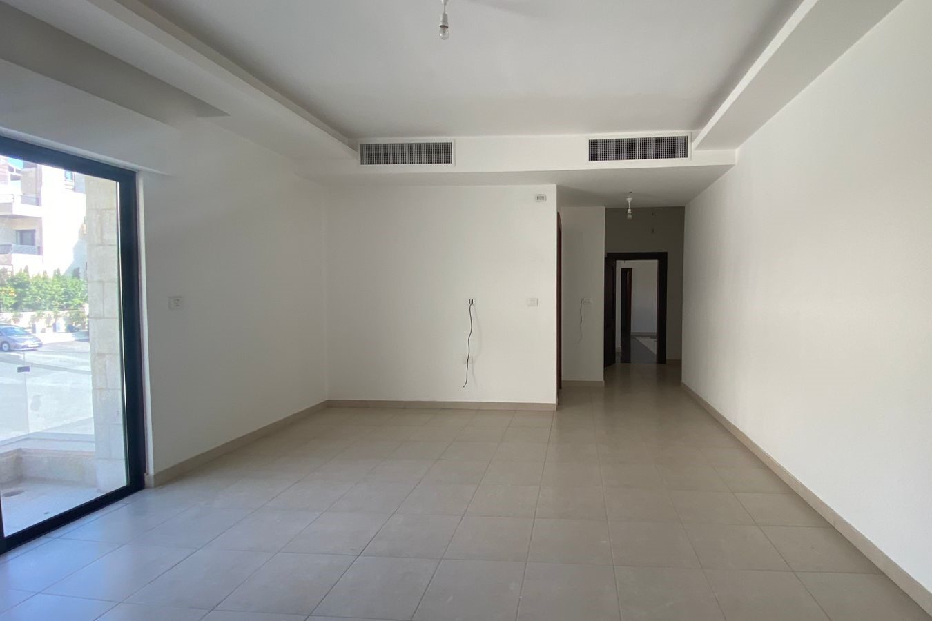 07efa46a-fa24-40b0-bafd-07fc257e4789 Empty, unfurnished interior with large glass door to street, white walls, and beige tile floor; hallway at far end with open doorways.