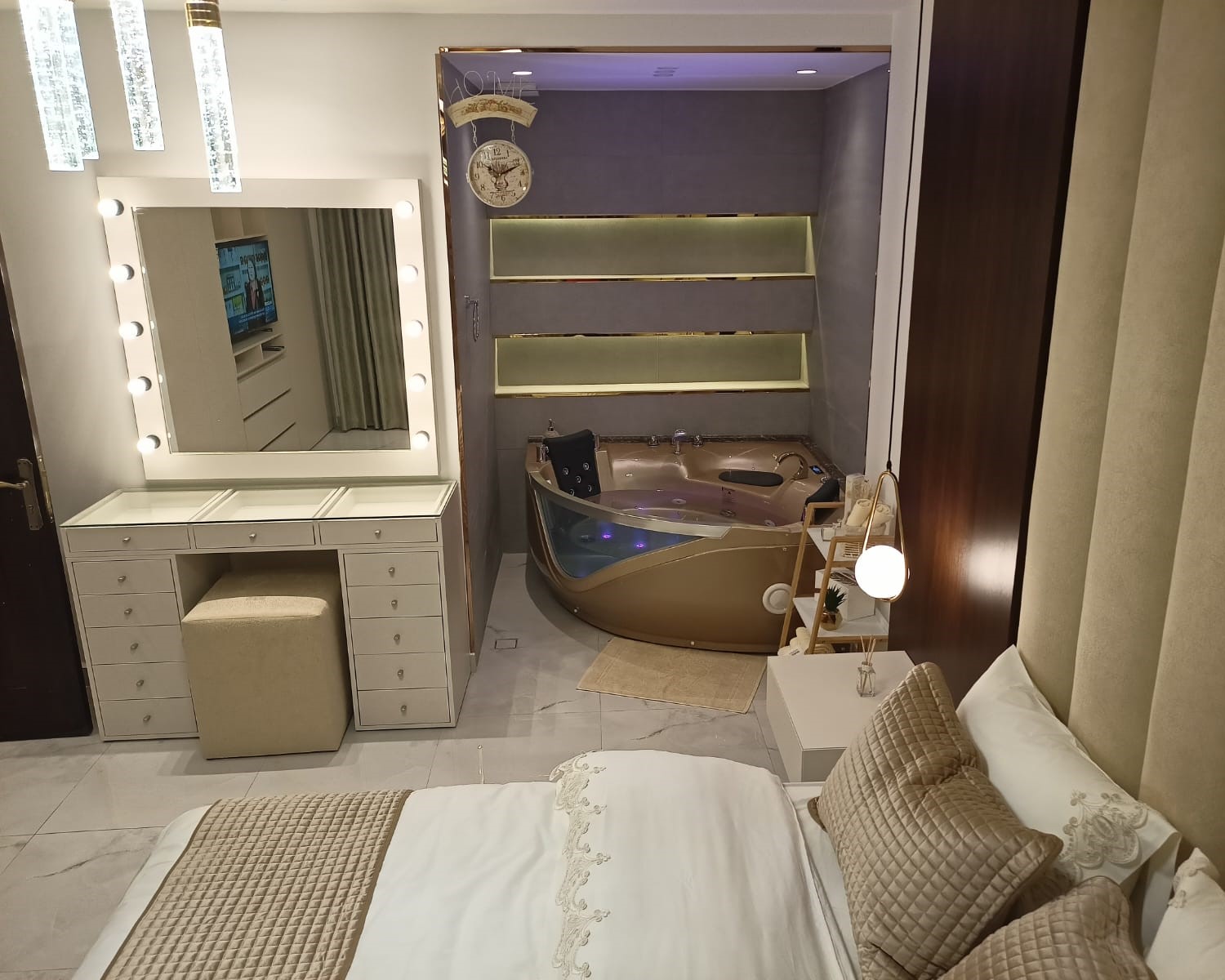 08a9810f-53b9-44ce-910e-74951c1f7506 Luxurious hotel bedroom with a lit vanity mirror, white dresser, and a beige stool, facing a glass-tenced bathtub area to the right.