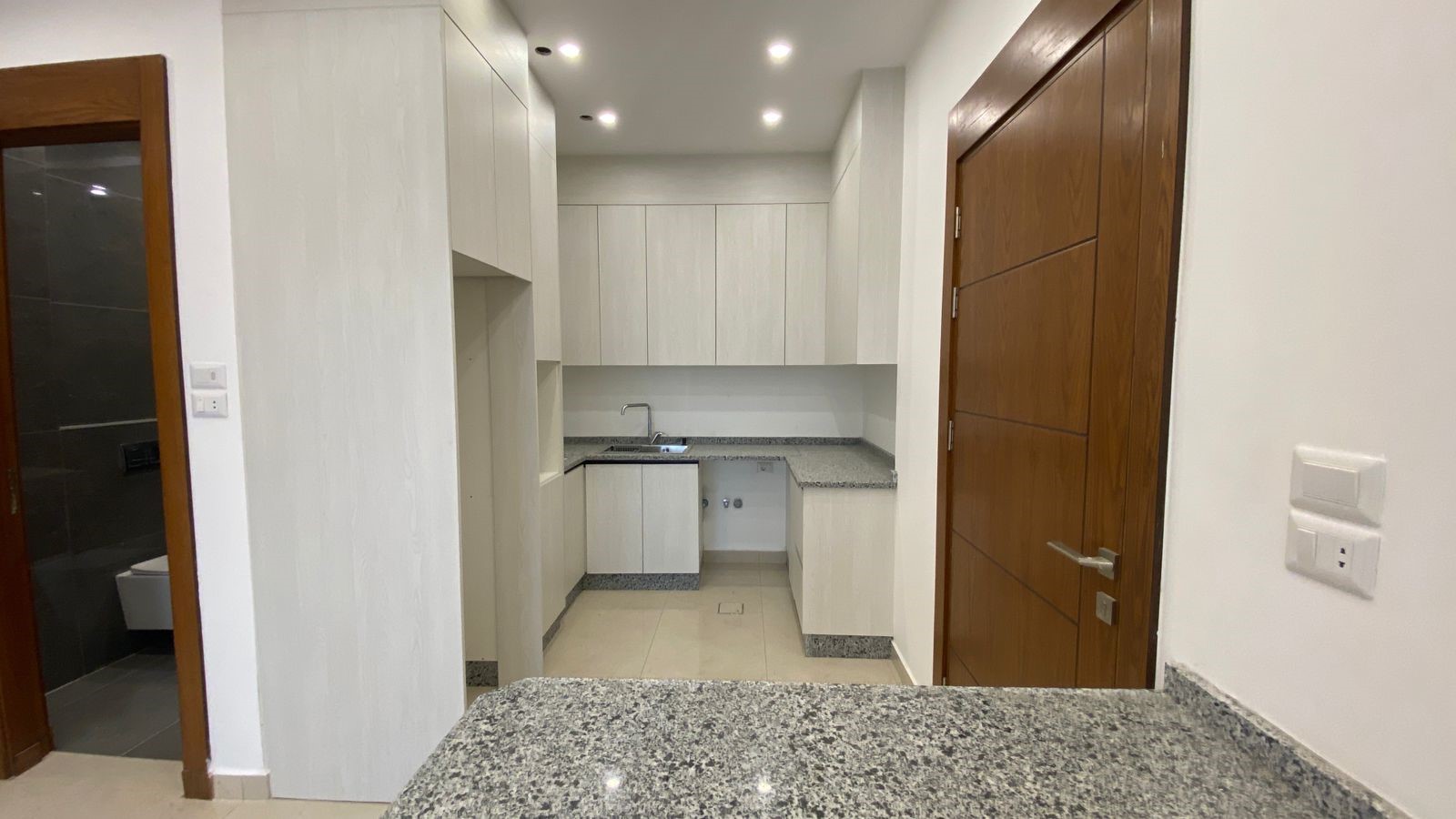 09907807-afe2-43a2-9862-ae6f3340f1e0 Modern white kitchen with granite countertops and stainless sink along the back wall, upper cabinets above and lower cabinetry below, adjacent doorway to a bathroom on the left and a wooden door on the right side.