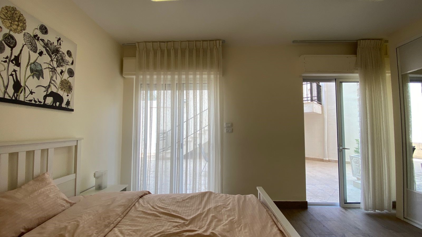 0a59e046-a775-42fe-9a3e-95a7b20f5e94 – Copy Bright bedroom with a white bed frame, pale pink bedding, sheer curtains, and a sliding glass door to a balcony.